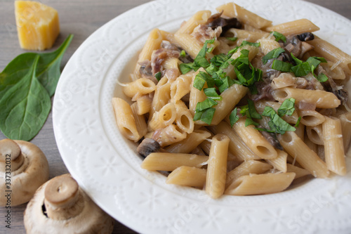 Vegan creamy mushroom garlic pasta with red onions garnished with basil in a white plate with its ingredients in the background, everything on the gray wooden surface