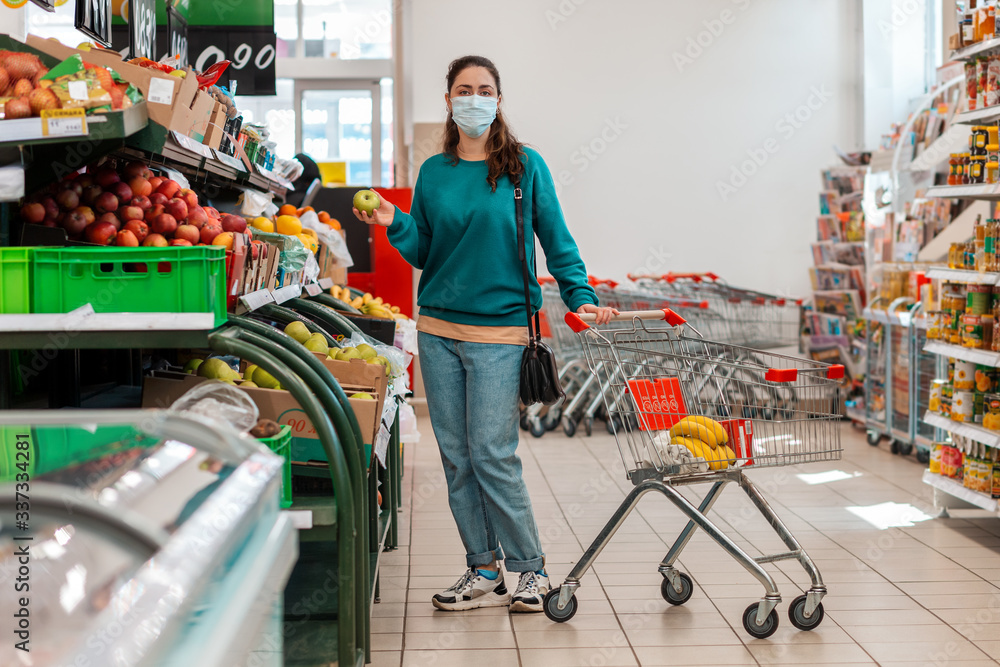 Shopping trip. A young Caucasian woman with a medical mask on her face holds a green apple in the vegetable Department of the store. The concept of buying products and pandemic coronavirus