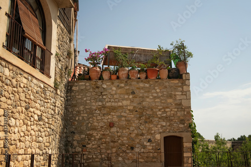 The wall of an old Spanish house is made of clay and stones with an arched window and an adjoining terrace with potted plants and flowers