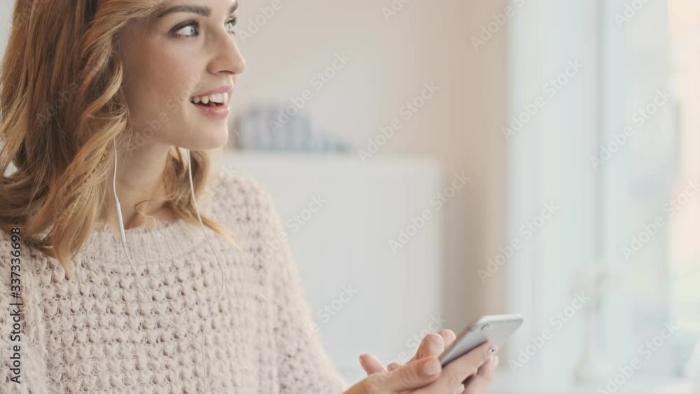 Lovely positive young blonde woman in wired earphones is using a mobile phone while drinking coffee in the white cozy cafe Lovely positive young blonde woman in wired earphones is using a mobile phone while drinking coffee in the white cozy cafe