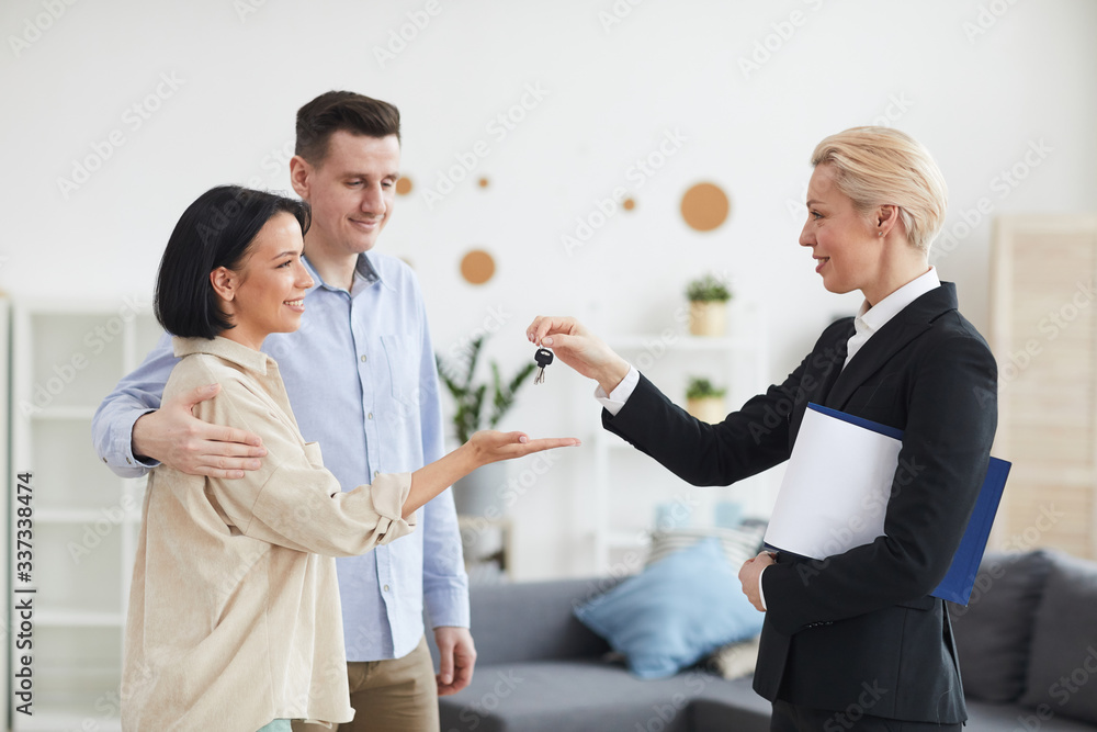 Mature real estate agent giving keys from new house to young couple while they standing in the living room at home Mature real estate agent giving keys from new house to young couple while they standing in the living room at home