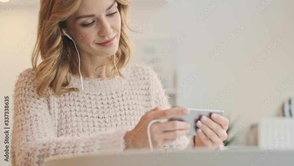 A positive calm woman using wired earphones is watching something on her smartphone while sitting in the white cozy cafe A positive calm woman using wired earphones is watching something on her smartphone while sitting in the white cozy cafe