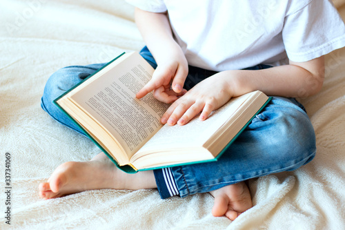 A child in blue jeans sits on the bed and holds an open book in his hands. Home leisure - reading a book.