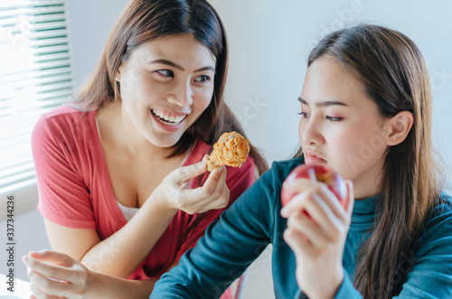 selective focus young woman smiling giving delicious hot fried chicken to friend holding red apple and refuse to eat sitting at home  weight loss  diet plan  healthy lifestyle and dieting food concept