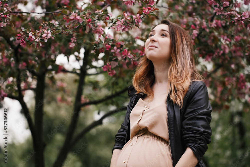 Young smilling pregnant woman looking to the sky wearing beige dress and black lather coat standing near blossoming tree in spring. horizontal portrait Young smilling pregnant woman looking to the sky wearing beige dress and black lather coat standing near blossoming tree in spring. horizontal portrait