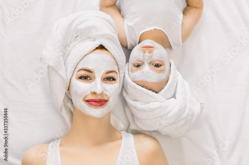 Portrait of pleasant young mother and her little daughter lying together on bed with white towels on head and facial clay mask while enjoying spa procedures. Isolated, top view. Beauty concept