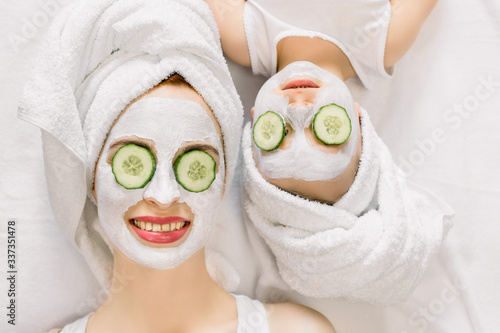Mother and daughter doing funny spa procedures after bath. They are in white bath towels with white facial mud mask on faces and slices of fresh cucumber on their eyes. Family spa, mothers day
