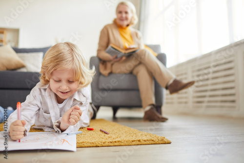 Little boy with blond hair lying on the floor and drawing with colored pencils with his grandmother reading a book in the background