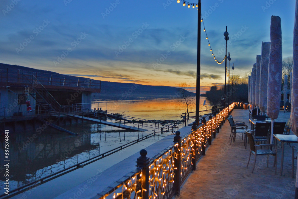 Obraz premium Landscape photography of restaurant terrace on the bank of the Danube river in Europe, evening colors of the sunset rays reflecting in the water with blue and orange hues