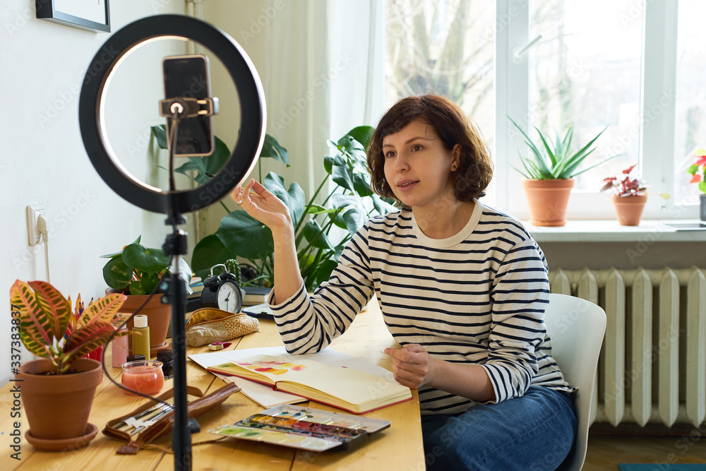 Female artist filming design workshop at home for online education. Working from home making master-class using ring lighting. Cozy artist's studio. Female artist filming design workshop at home for online education. Working from home making master-class using ring lighting. Cozy artist's studio.