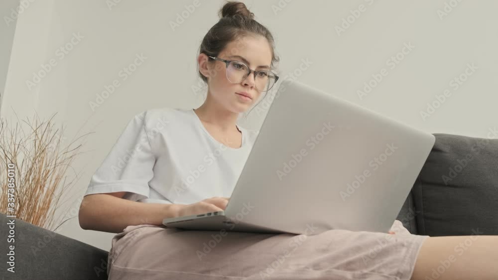 View from below of Concentrated pretty brunette woman in eyeglasses using laptop computer while sitting on sofa at home View from below of Concentrated pretty brunette woman in eyeglasses using laptop computer while sitting on sofa at home