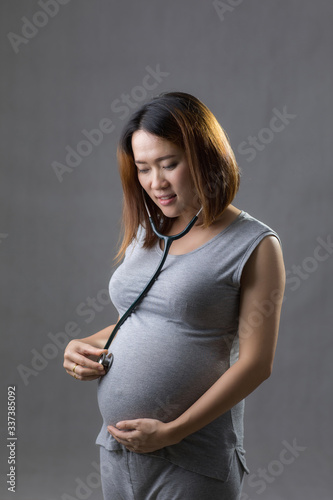 Pregnant woman in gray dress hold hands on belly with stethoscope in white background.
