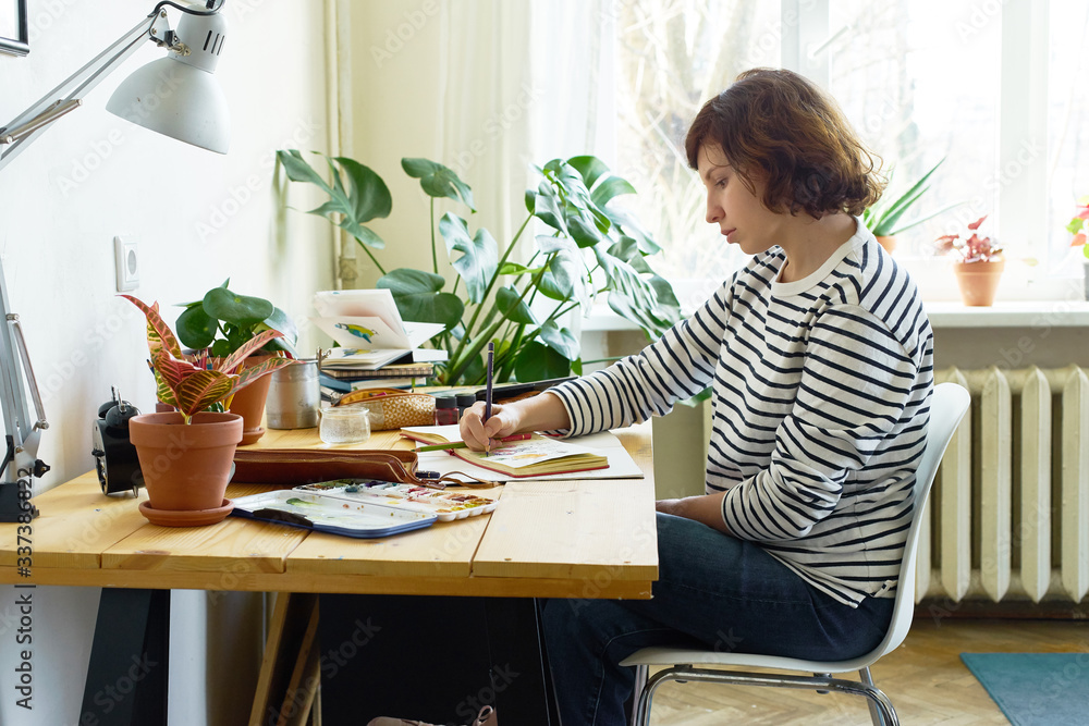 Female artist at her workplace working from home. Woman dressed in jeans and striped shirt sitting at the table and drawing illustration. Female artist at her workplace working from home. Woman dressed in jeans and striped shirt sitting at the table and drawing illustration.