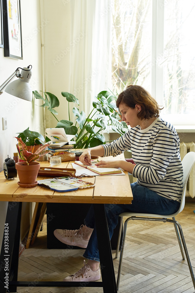 Female artist at her workplace working from home. Woman dressed in jeans and striped shirt sitting at the table and drawing illustration.