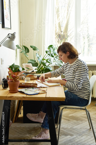 Female artist at her workplace working from home. Woman dressed in jeans and striped shirt sitting at the table and drawing illustration.