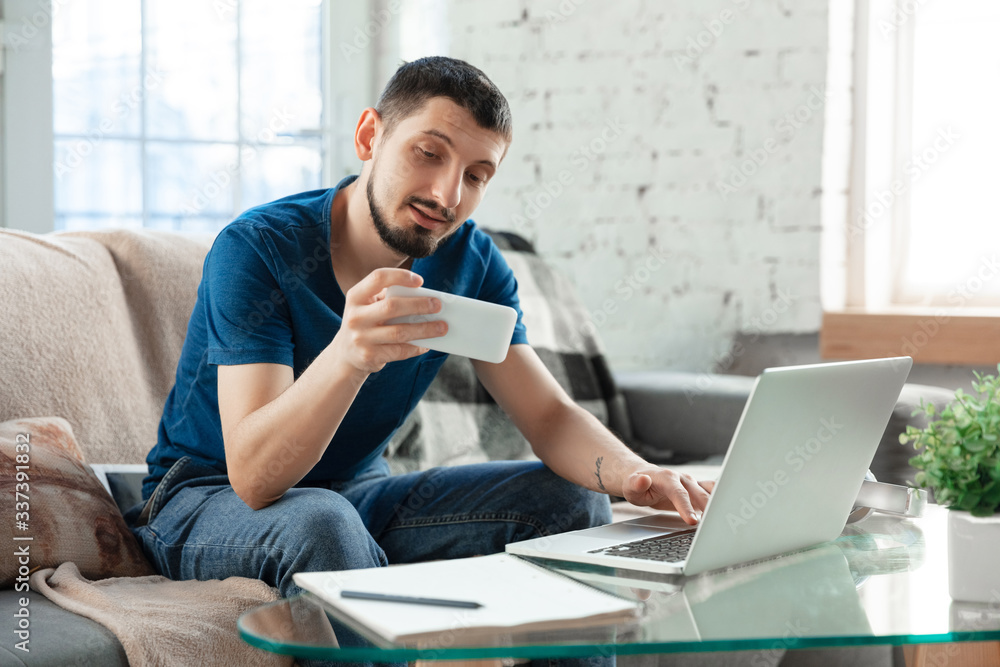 Young focused man studying at home during online courses or free information by hisself. Becomes economist, financist, translator while isolated, quarantined. Using laptop, smartphone, headphones. Young focused man studying at home during online courses or free information by hisself. Becomes economist, financist, translator while isolated, quarantined. Using laptop, smartphone, headphones.