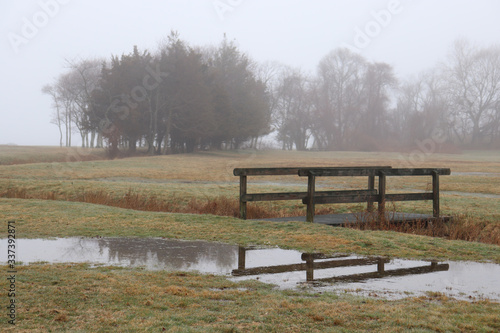 Small wooden bridge over ditch with reflection in rain puddle and trees in background covered in light fog