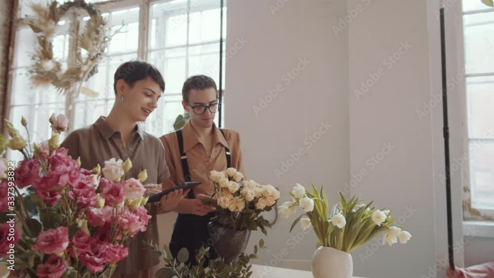 Tilt down shot of professional male florist in apron telling about roses to young woman while she using digital tablet in flower shop Tilt down shot of professional male florist in apron telling about roses to young woman while she using digital tablet in flower shop