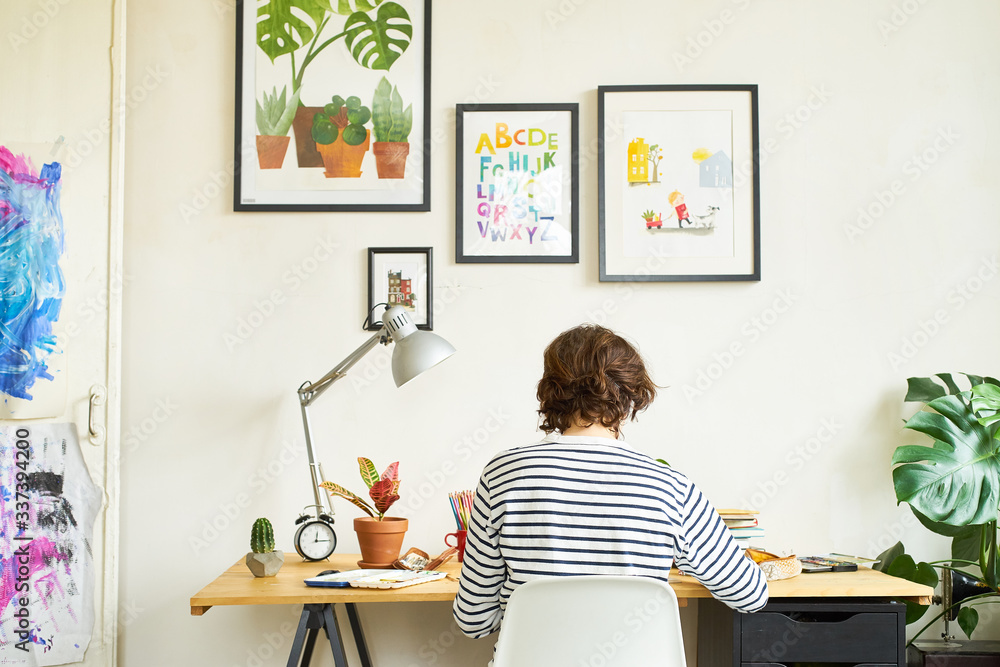 Female artist at her workplace working from home. Young woman dressed in jeans and striped shirt sitting at the table turned backwards. Creating an illustration. Female artist at her workplace working from home. Young woman dressed in jeans and striped shirt sitting at the table turned backwards. Creating an illustration.