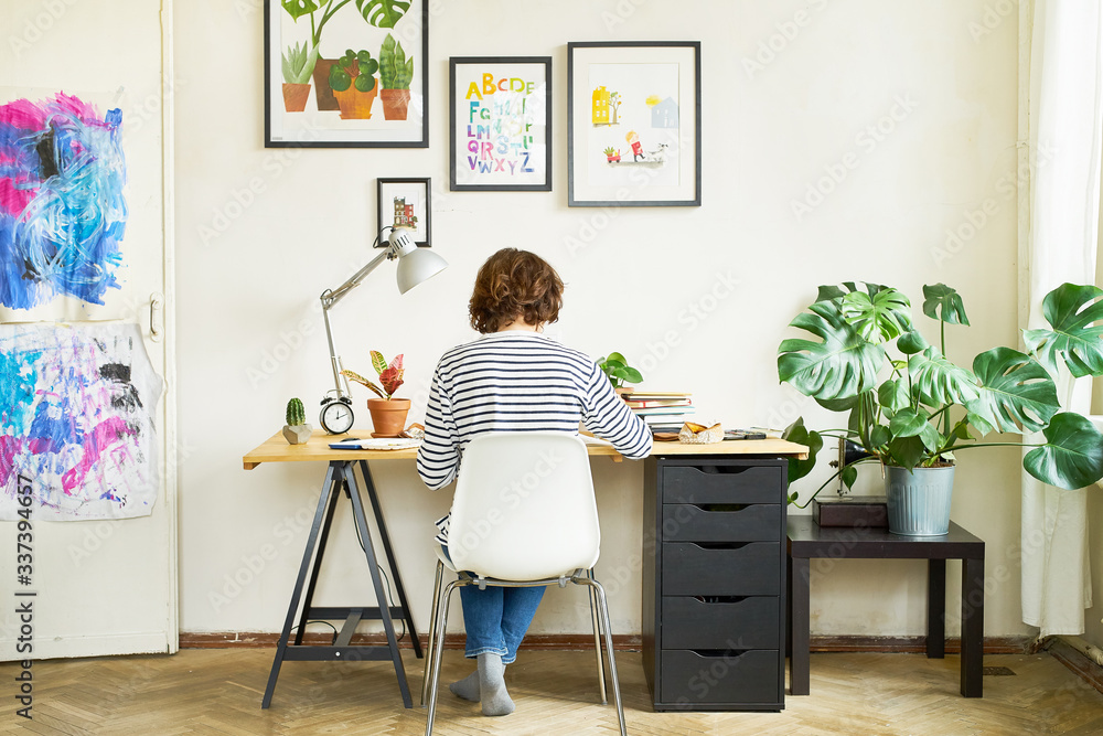 Fototapeta premium Female artist at her workplace working from home. Young woman dressed in jeans and striped shirt sitting at the table turned backwards. Creating an illustration.