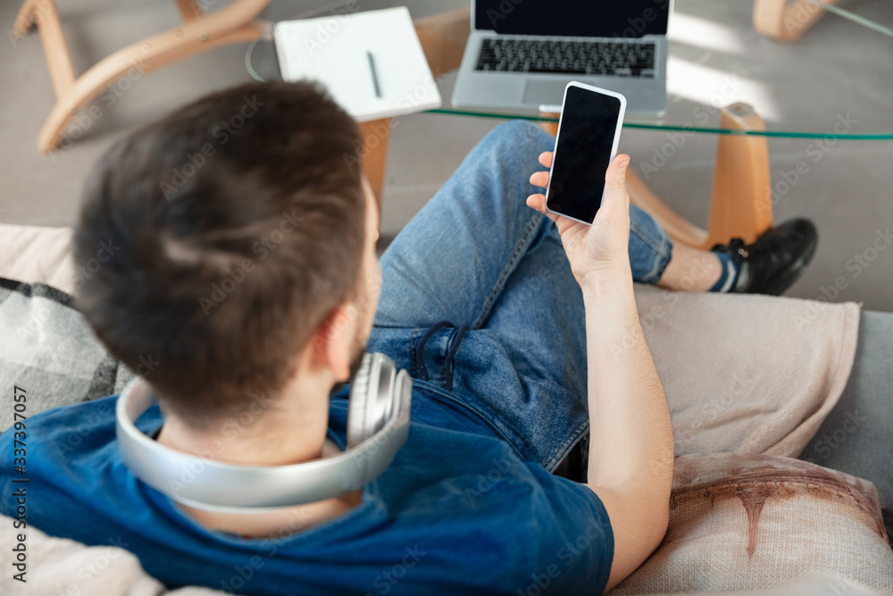 Young focused man studying at home during online courses or free information by hisself. Becomes nurse, constraction laborer while isolated, quarantined. Using laptop, smartphone, headphones. Young focused man studying at home during online courses or free information by hisself. Becomes nurse, constraction laborer while isolated, quarantined. Using laptop, smartphone, headphones.