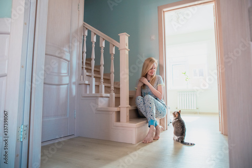 blonde girl sits on the bottom step of the stairs in a house with a bright interior and looks at the cat