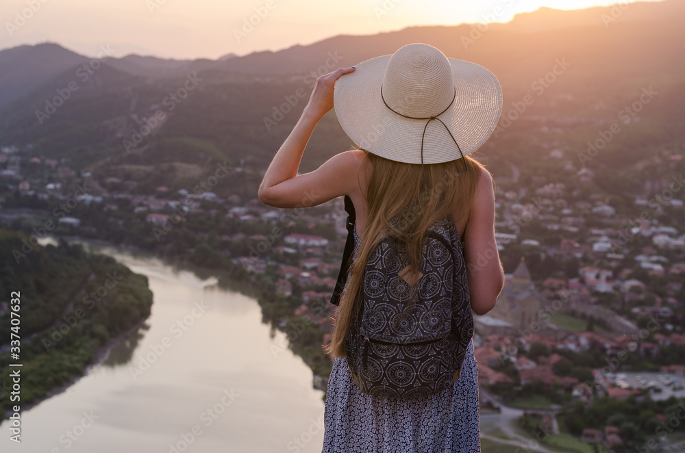 Young beautiful girl in dress and hat, looking at river and city sunset.