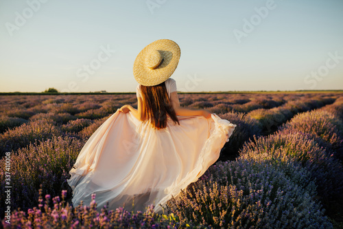 Beautiful young woman walking the field of lavender in Provence, France. Fashion outfit pale rose dress, straw hat. Back view. Violet in nature. Back view of beautiful girl in a dress and straw hat.