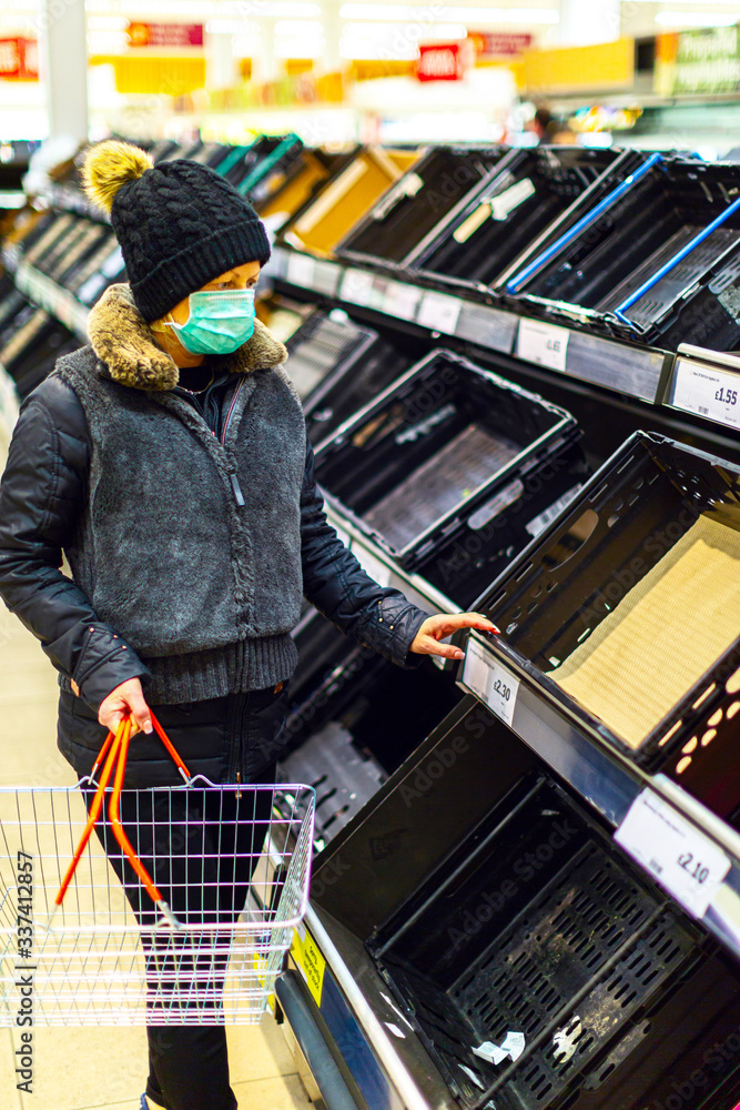 Female customer wearing a protective medical mask looks confused at the empty shelves of fruits and vegetables in a supermarket