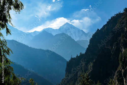 View on Himalayan valley along Annapurna Circuit Trek, Nepal. There is a dense forest in front. High, snow caped mountains' peaks catching the sunbeams. Serenity and calmness. Barren slopes