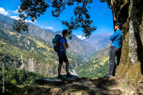 A couple in hiking outfits standing under a thick tree trunk and enjoying the view on lush green Himalayan valley along Annapurna Circuit in Nepal. They are happy and full of energy. Cheerful moments photo