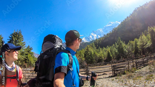 A couple taking a selfie while trekking along Annapurna Circuit in Nepal. They are having fun and making silly faces. There is a lush green Himalayan valley around. Snow caped mountains in the back