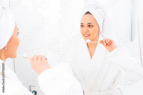 Girl with a toothbrush. Young woman brushes her teeth looking in the mirror in the bathroom. Photo of a woman in a bathrobe in the bathroom.