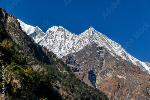 A close up view on snow caped Himalayan peak seen from Annapurna Circuit Trek, Nepal. Sharp and steep slopes of the mountain. Lower parts of the mountain overgrown with some plants. Harsh landscape. © Chris