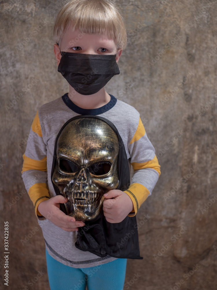 Funny blond kid with scary skeleton mask in his hands in black medical mask to protect against influenza viruses in crowded places. Chinese inscription on the wall background, Wuhan coronavirus