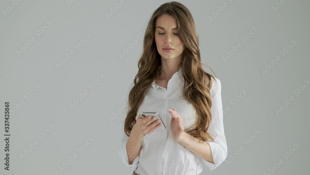 A beautiful woman is waving hand to someone while using her smartphone isolated over a gray background in studio A beautiful woman is waving hand to someone while using her smartphone isolated over a gray background in studio