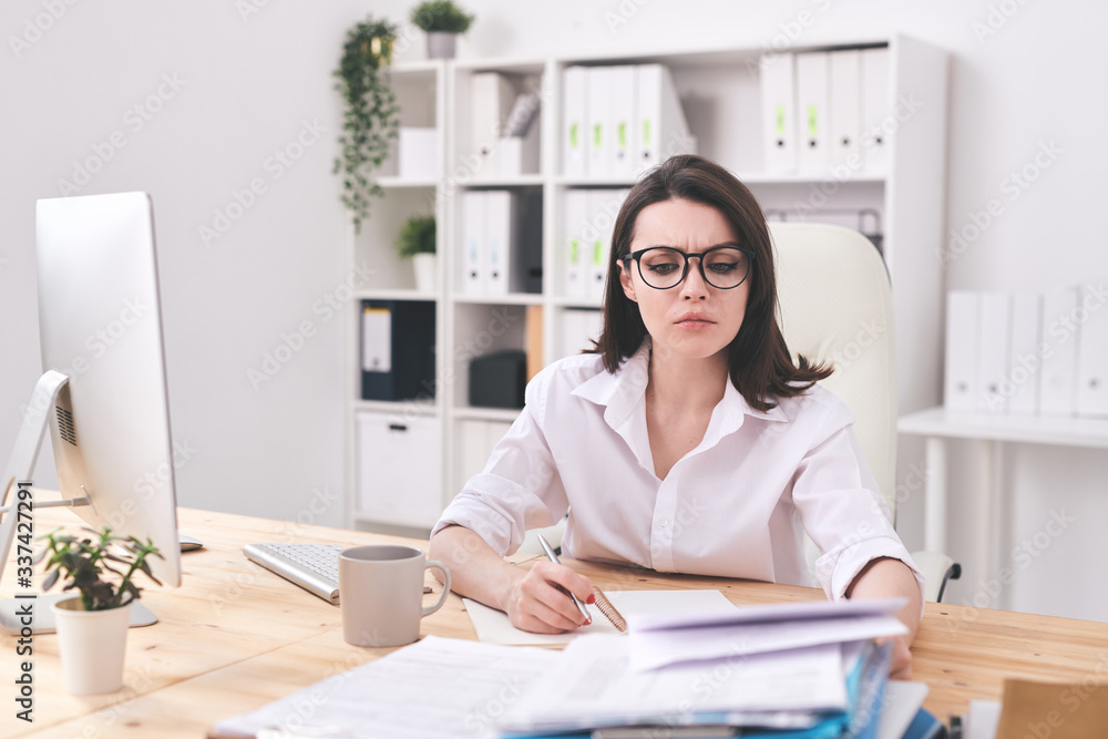 Young serious businesswoman looking at financial paper while making notes Young serious businesswoman looking at financial paper while making notes