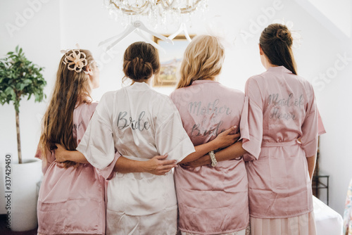 The bride and bridesmaids in silk robes of white and pink color stand with their backs to the camera and hug each other at the waist