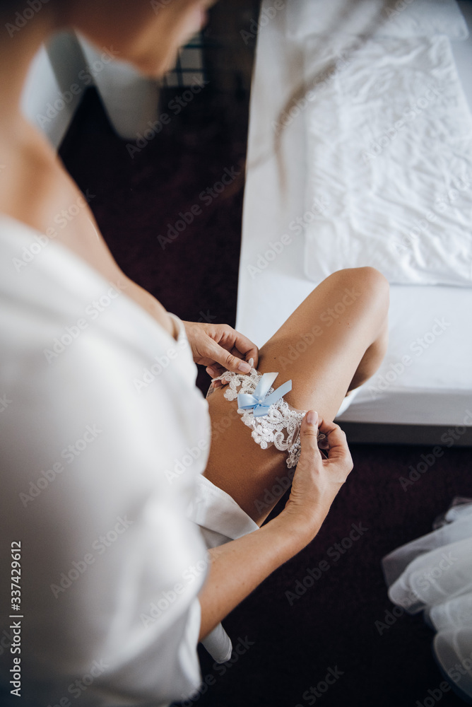 The bride puts on a garter on her leg, white with a blue bow, embroidered with beads The bride puts on a garter on her leg, white with a blue bow, embroidered with beads