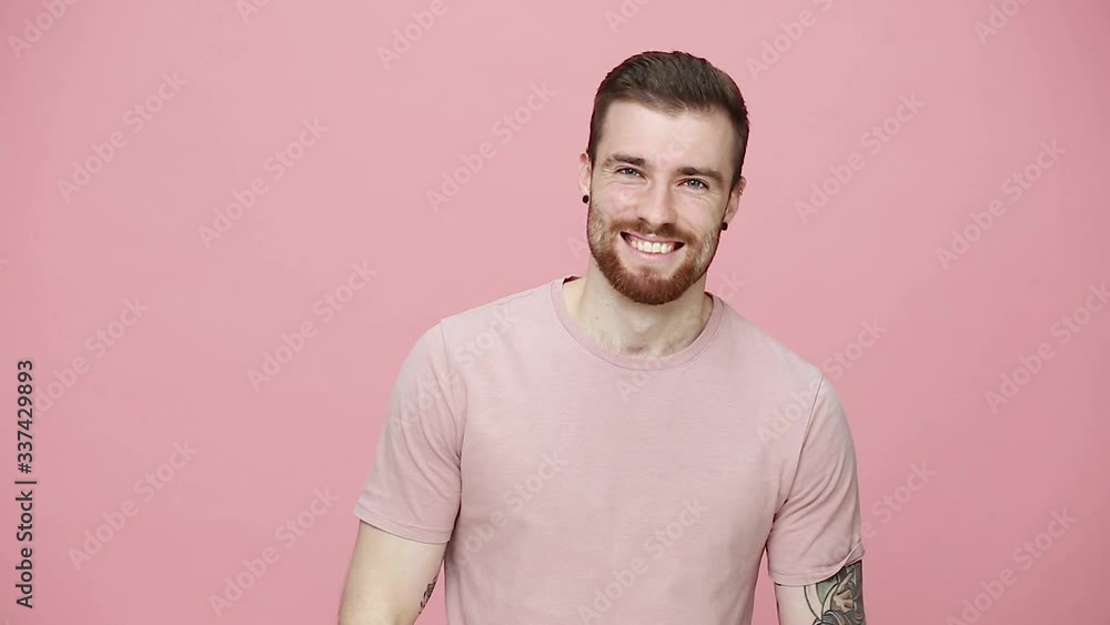 Handsome bearded tattooed young guy 20s in t-shirt isolated on pastel pink background studio. People lifestyle concept. Look at camera with charming smile waving greeting with hand as notices someone Handsome bearded tattooed young guy 20s in t-shirt isolated on pastel pink background studio. People lifestyle concept. Look at camera with charming smile waving greeting with hand as notices someone