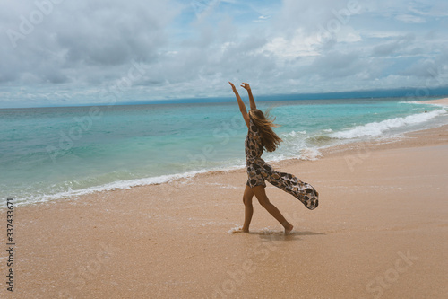 Beautiful and young girl in a dress posing on the shore of the ocean and sea with blue waves. Woman in a long dress model and fashion posing. Relax and relaxation on the island of Bali