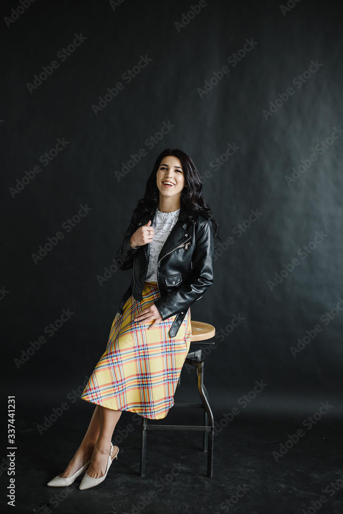 portrait of a young dark-haired girl on a dark background sitting on a bar stool with dark leather jacket and skirt. Photography of a young beautiful girl in the Studio portrait of a young dark-haired girl on a dark background sitting on a bar stool with dark leather jacket and skirt. Photography of a young beautiful girl in the Studio