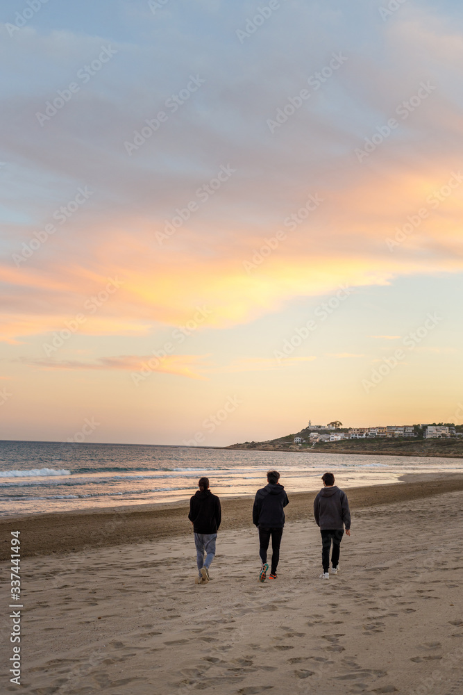 Three young boys walk on the beach towards the sunset