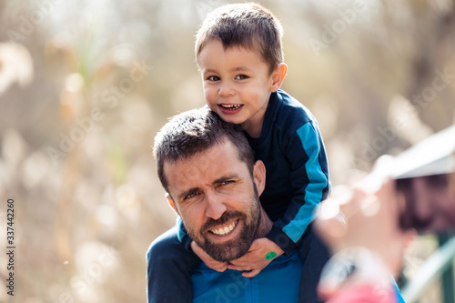 Handsome young father with his little son on the shoulders looking at the camera while they take a photo outdoors.