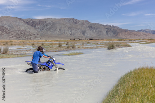 A motorcyclist takes a motorcycle through a white mountain river, his friend helps him. Mongolia, Altai.