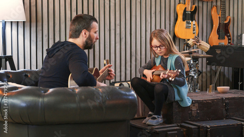 Dad teaching guitar and ukulele to his daughter.Little girl learning guitar at home.Side view.Ukulele class at home. Child learning guitar from her father