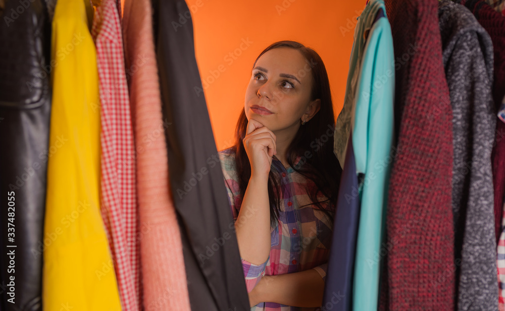 Young woman grimacing standing between clothes in wardrobe. Adult female choosing things in store.