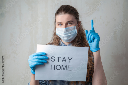 A young girl in a medical mask and gloves with an inscription on a sheet of paper calls for staying in quarantine at home. Home self-isolation in pandemic, flu epidemic, viral diseases, Covid-19.