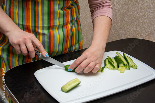 closeup hands girl in a striped apron in the kitchen is slicing cucumbers into strips on a whiteBoard on the black table.