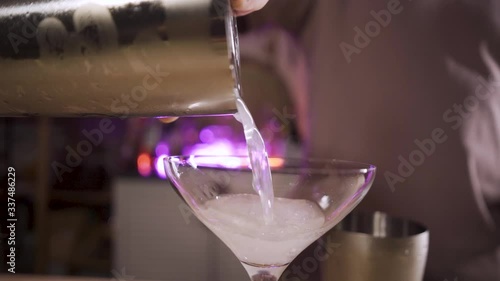 Close-up shot of the male hand pouring a cocktail from a shaker with a strainer into a glass. The liquid flows beautifully against a background of purple light. Slow motion.